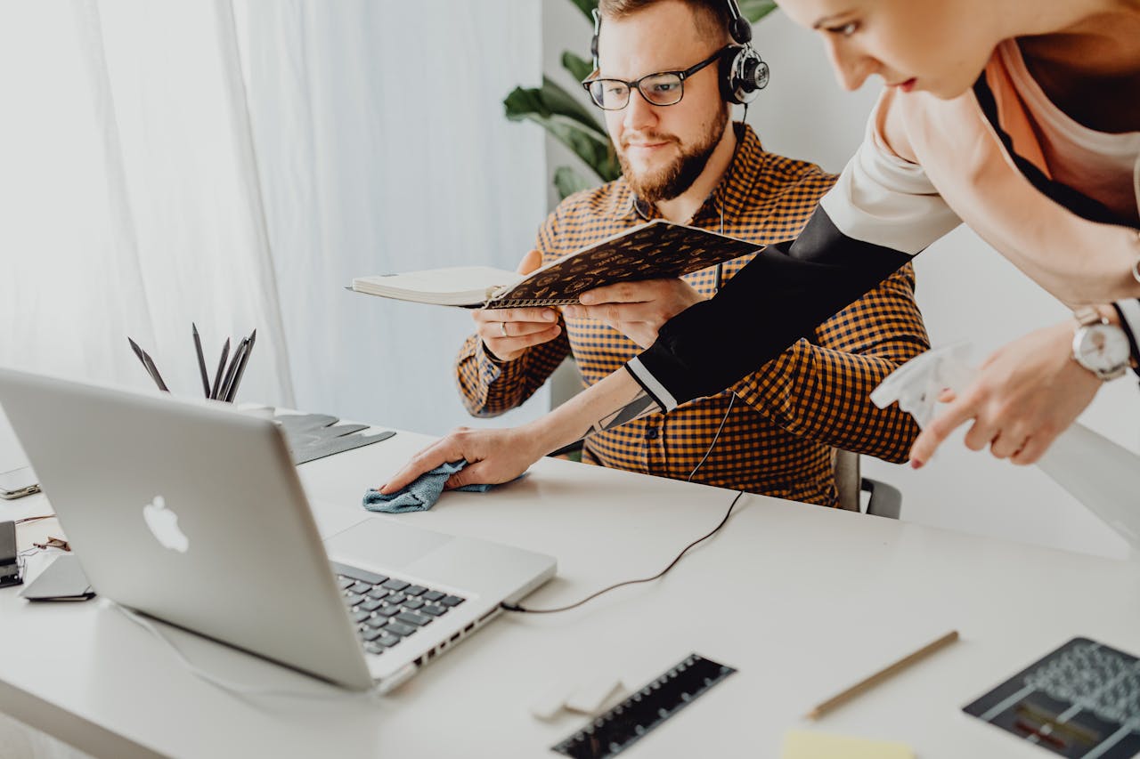 A man working remotely while a woman cleans the desk with a spray.